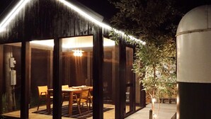 Una casa moderna con una terraza de madera, una zona de comedor con sillas y un depósito metálico grande.