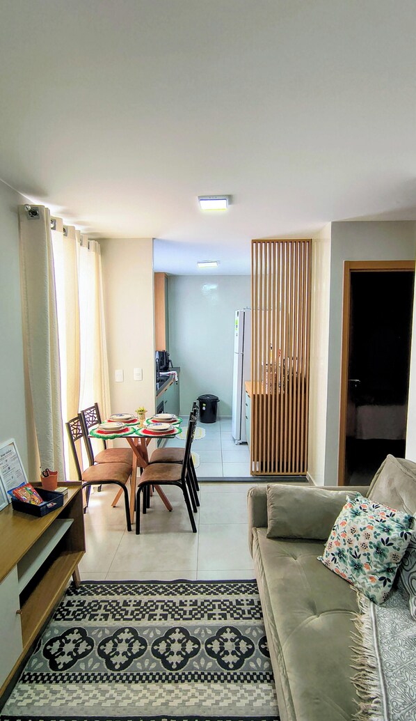 Uma sala de estar com sofá, mesa de jantar e cadeiras, cozinha e uma porta deslizante que dá para outro quarto.