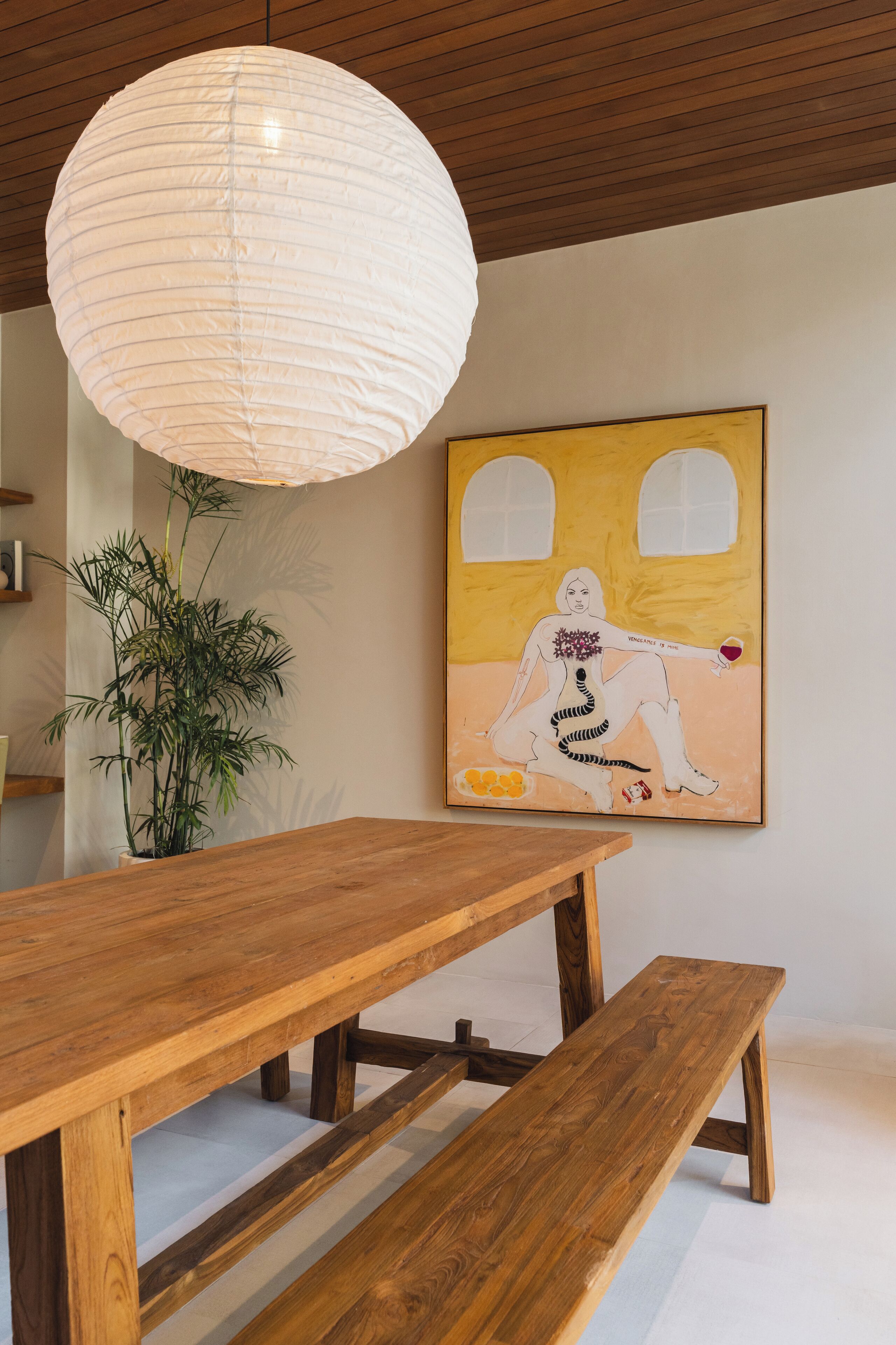 A wooden dining table with benches, a hanging paper lantern, a potted plant, and a painting on the wall.