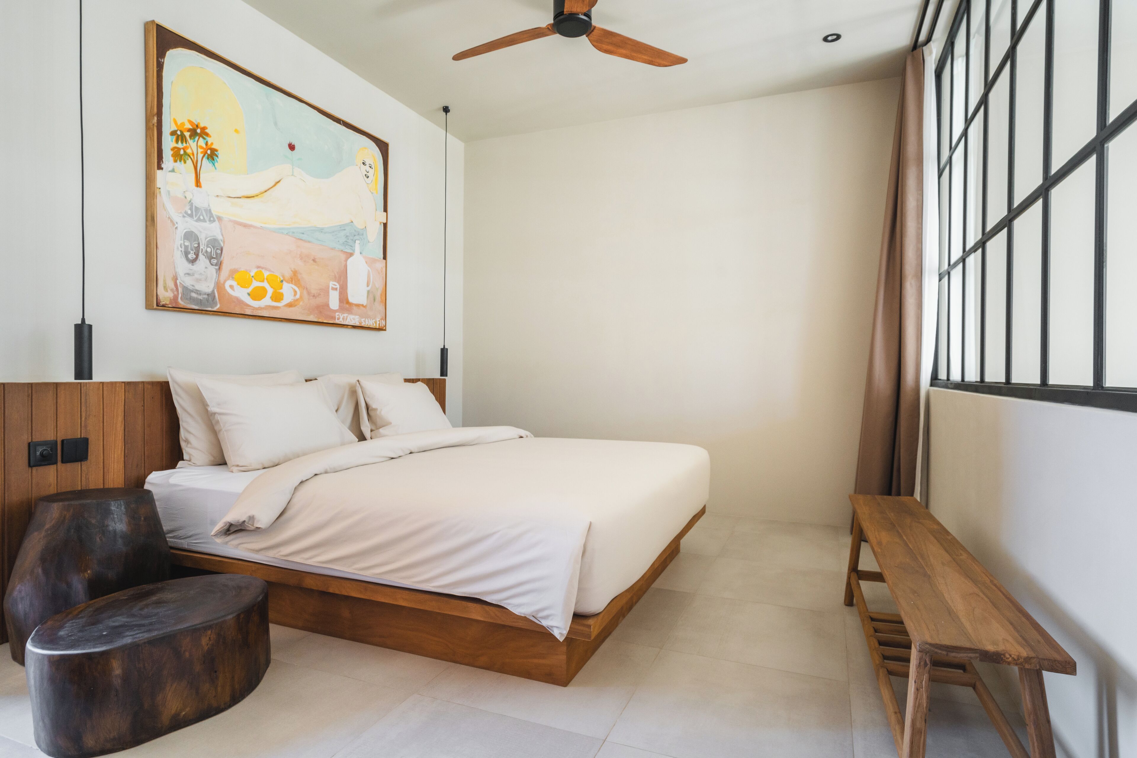 Une chambre à coucher moderne avec un lit en bois, un banc, une œuvre d’art murale originale et un ventilateur de plafond.