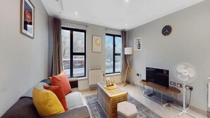 A modern living room with a sofa, a TV, a fan, and a window with curtains. - Apartment - Sleeps 2 - Near Tube Stn (London)