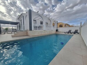 A swimming pool with a white building in the background, a covered area with benches, and a clear sky.