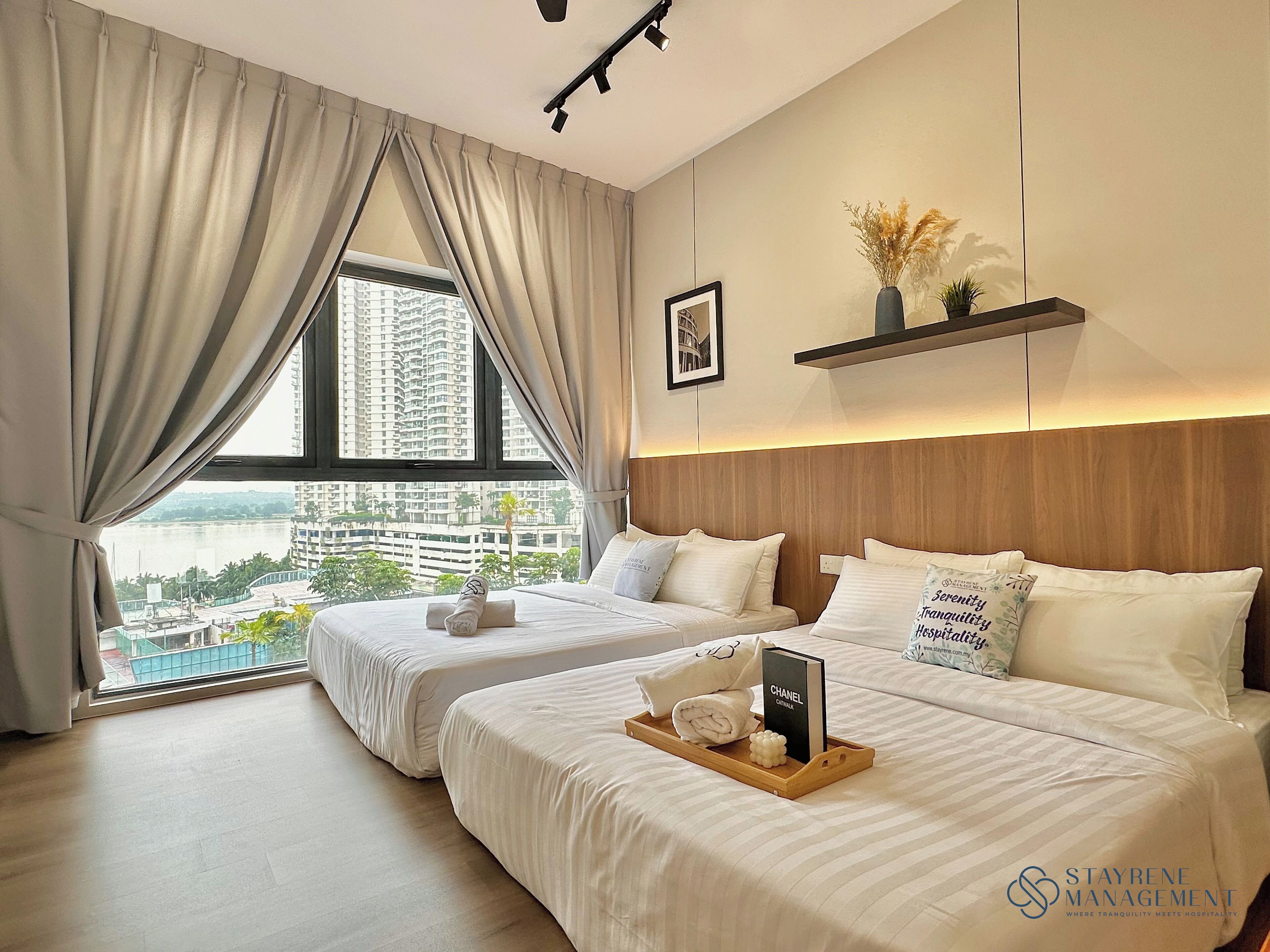 A hotel room with two beds, a large window with curtains, a view of a cityscape, a framed picture on the wall, and a tray with a book and towels.
