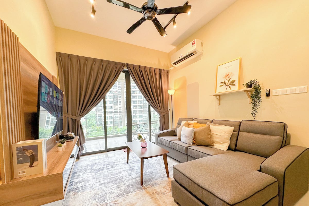A modern living room with a sofa, a coffee table, and a TV. There is a large window with curtains, an air conditioning unit, and a ceiling fan.
