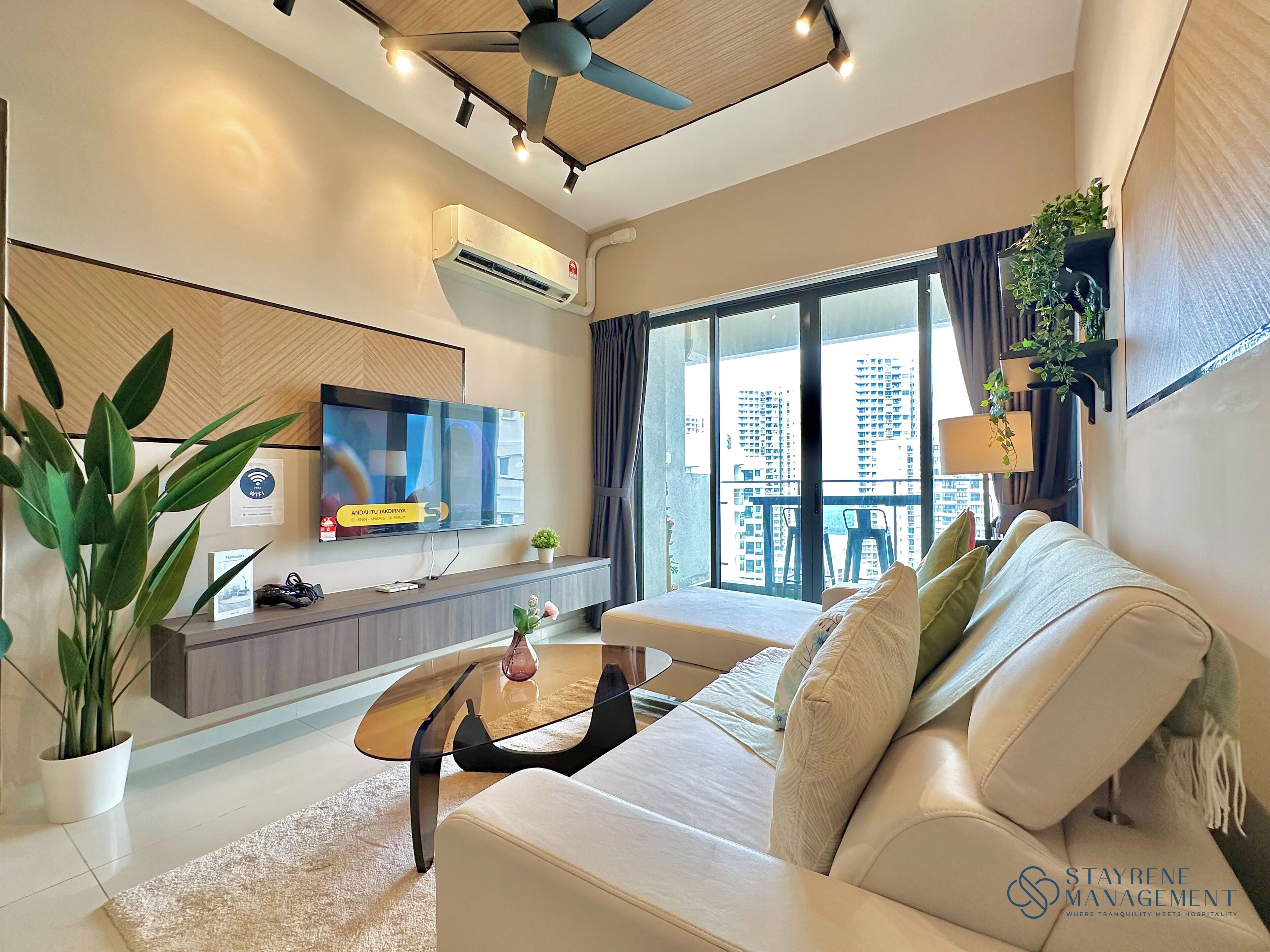 A modern living room with a sofa, coffee table, and a flat-screen TV. There is a balcony with a view of high-rise buildings.