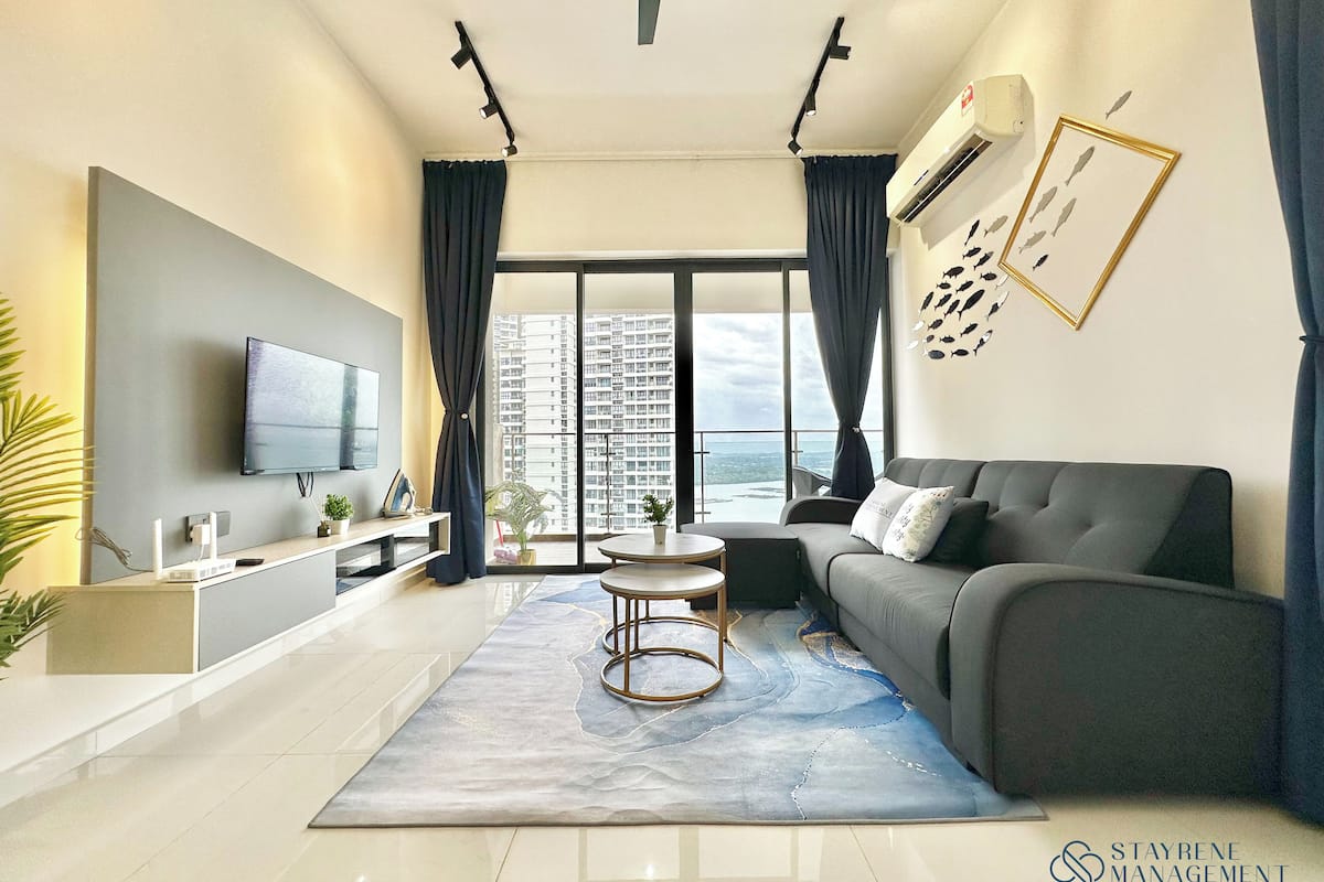 A modern living room with a flat-screen TV, a grey sofa, a coffee table, and a view of high-rise buildings.