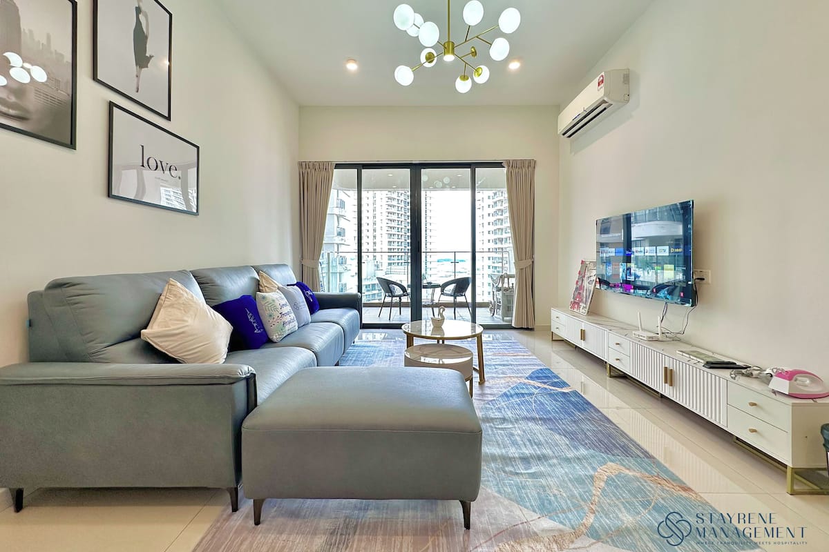 A modern living room with a grey sofa, a matching ottoman, and a large TV. There is a balcony with a view of high-rise buildings.