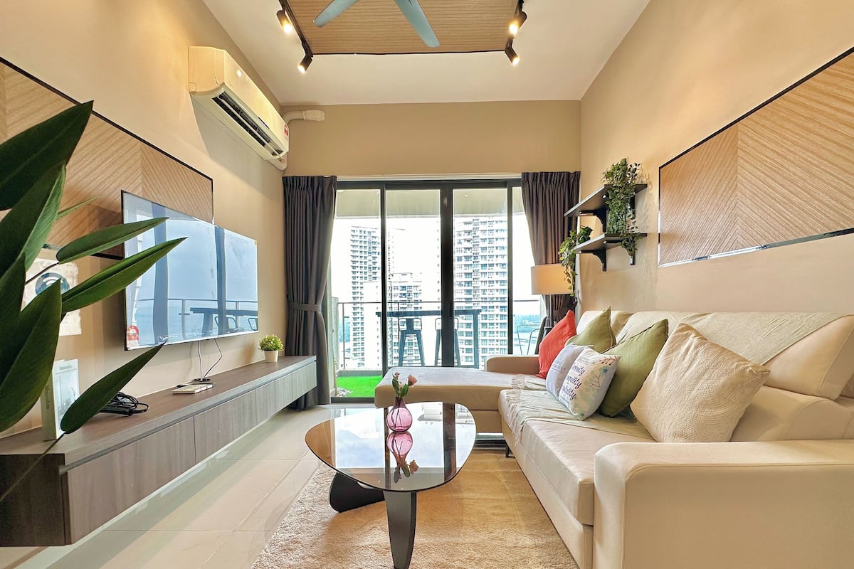 A modern living room with a sofa, a coffee table, and a TV. There is a large window with a view of buildings.
