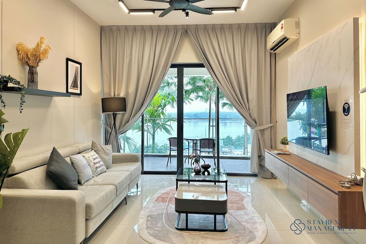 A modern living room with a sofa, coffee table, and TV. There is a balcony with a view of palm trees and a body of water.