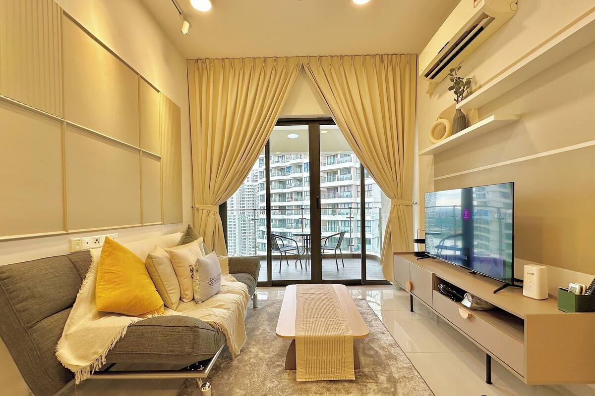 A modern living room with a sofa, a coffee table, and a TV. There is a balcony with a view of high-rise buildings.