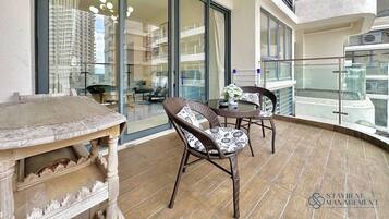 A balcony with a table and two wicker chairs, a glass railing, and a view of a high-rise building.