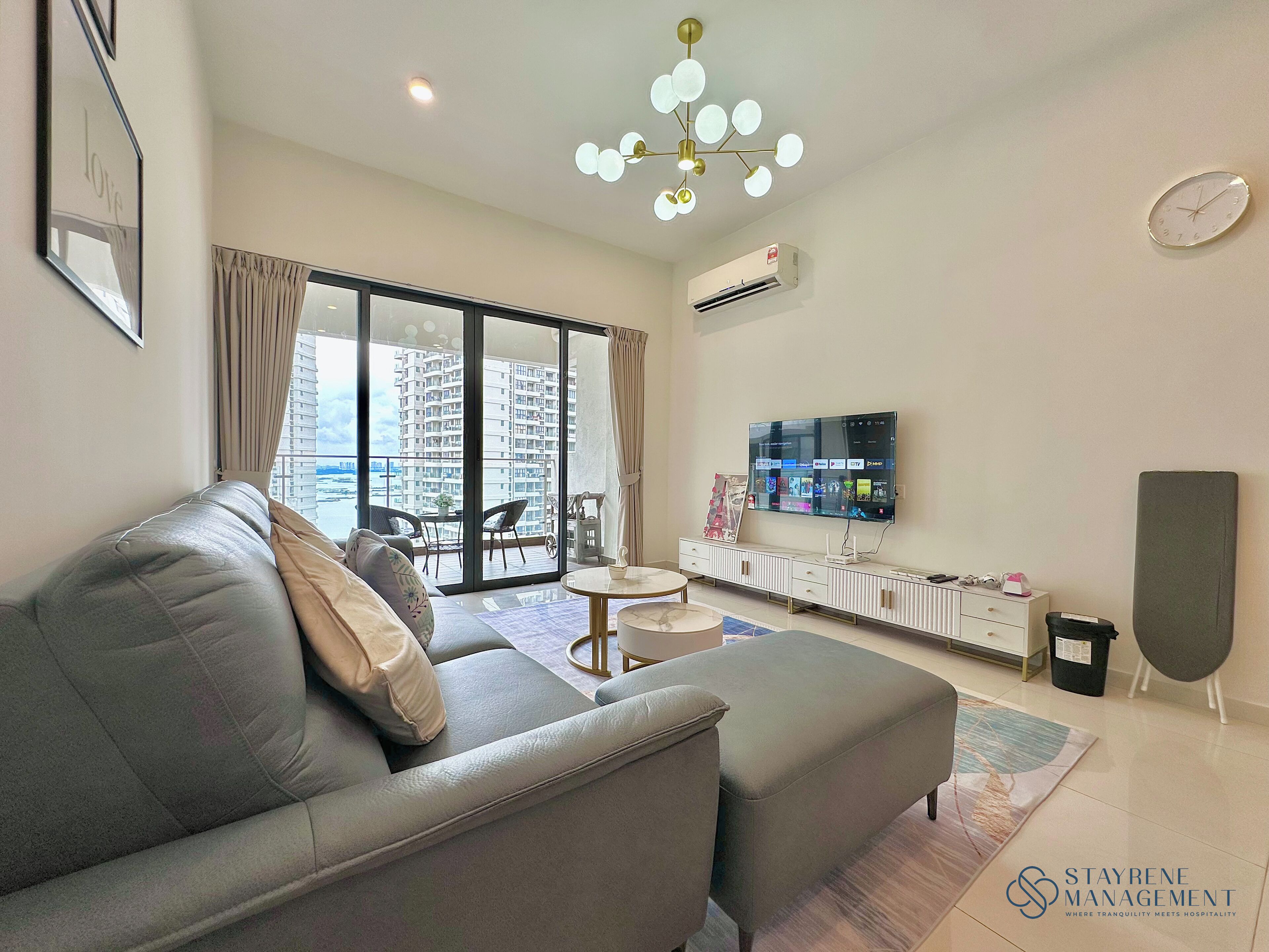A modern living room with a grey sofa, a flat-screen TV, a ceiling light fixture, and a view of a high-rise building.