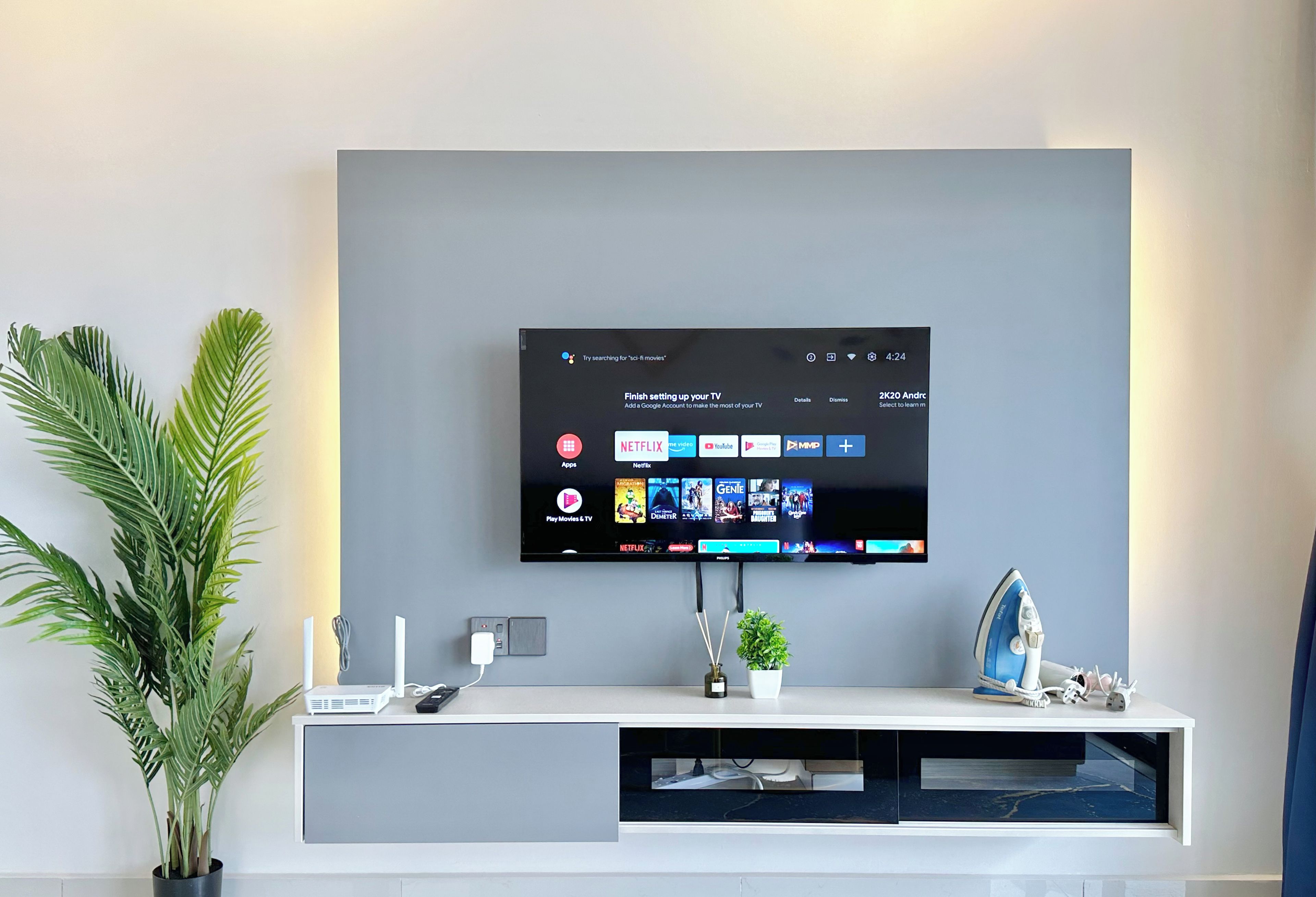 Una sala de estar moderna con un televisor montado que muestra aplicaciones de transmisión, un mueble blanco para entretenimiento y una planta en maceta.