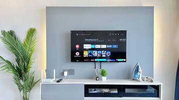 Una sala de estar moderna con un televisor montado que muestra aplicaciones de transmisión, un mueble blanco para entretenimiento y una planta en maceta.