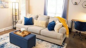 Una sala de estar con un sofá, un reposapiés y un sillón azul. Hay una bandeja con un florero y un control remoto sobre el reposapiés.