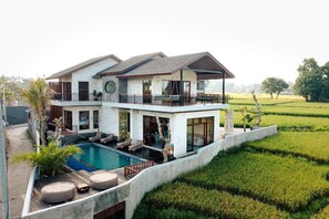 Una casa de dos pisos con piscina, mobiliario exterior y vistas a un campo de arroz.