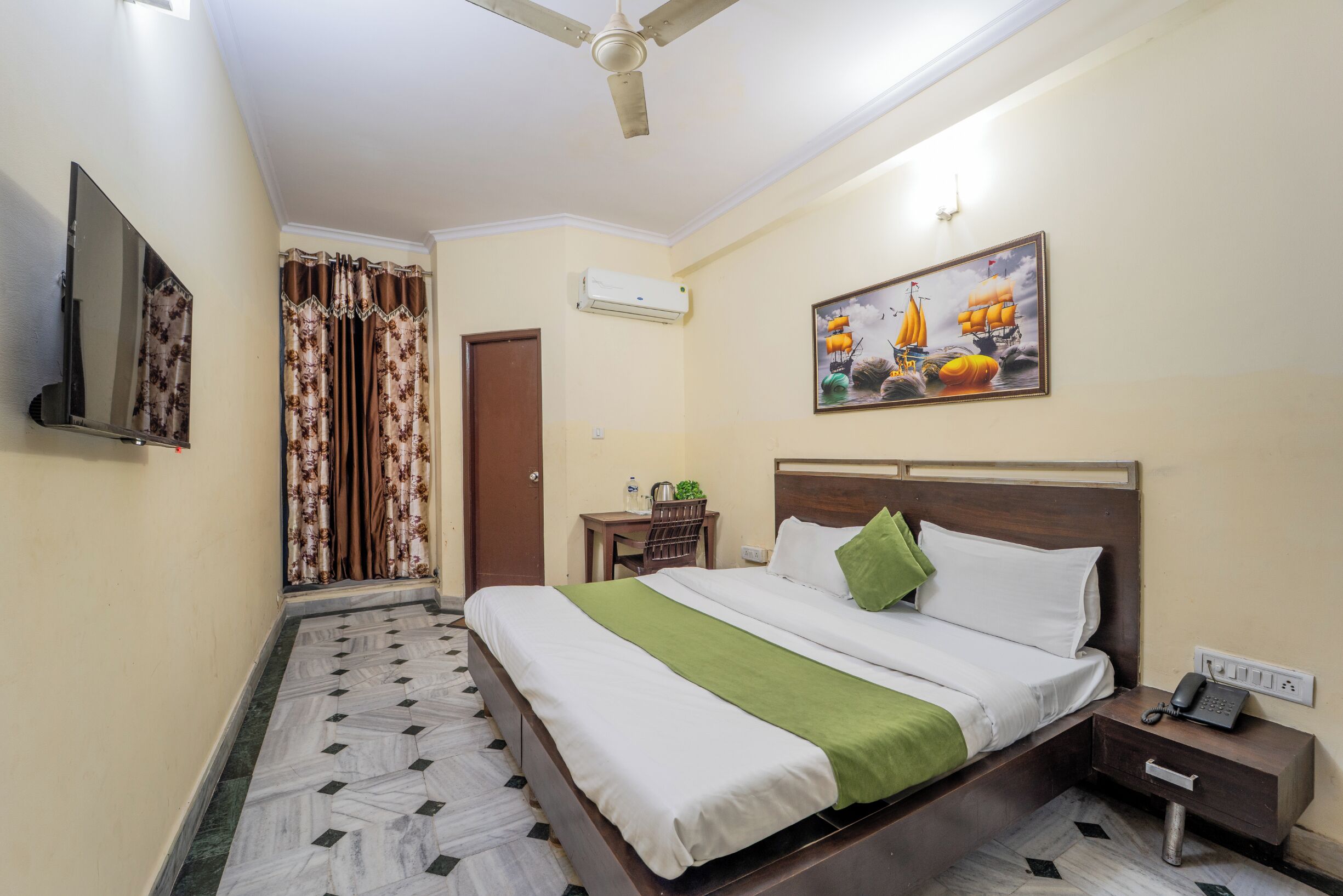 A hotel room with a bed, bedside table, nightstand, TV, air conditioning unit, and a painting on the wall.