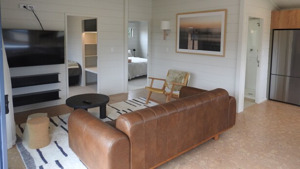 A modern living room with a brown leather sofa, a black coffee table, and a patterned rug. There is a television mounted on the wall, a framed picture above the sofa, and a kitchen area visible in the background. - Hinenamu Cottage -Two Bedroom and located in a quiet Cul de Sac! (Turangi)