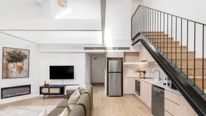 A modern living room with a sofa, a flat-screen TV, and a fireplace. There is a kitchen with a refrigerator, stove, and sink. A staircase with a black railing leads to an upper level. - The Henrietta Apartments by Urban Rest (Double Bay)