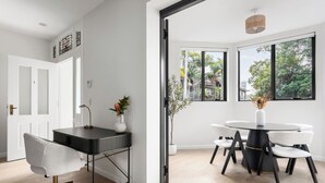 A modern room with a desk, chair, and round table with chairs. There are windows with a view of trees and buildings. - The Henrietta Apartments by Urban Rest (Double Bay)