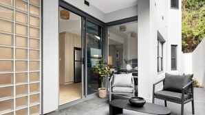 A modern patio area with a sliding glass door leading to a kitchen, a round table, two chairs, and a potted plant. - The Henrietta Apartments by Urban Rest (Double Bay)