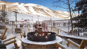 Un área de estar al aire libre cubierta de nieve, con un pozo de fuego, sillas de madera y vistas a una montaña nevada.