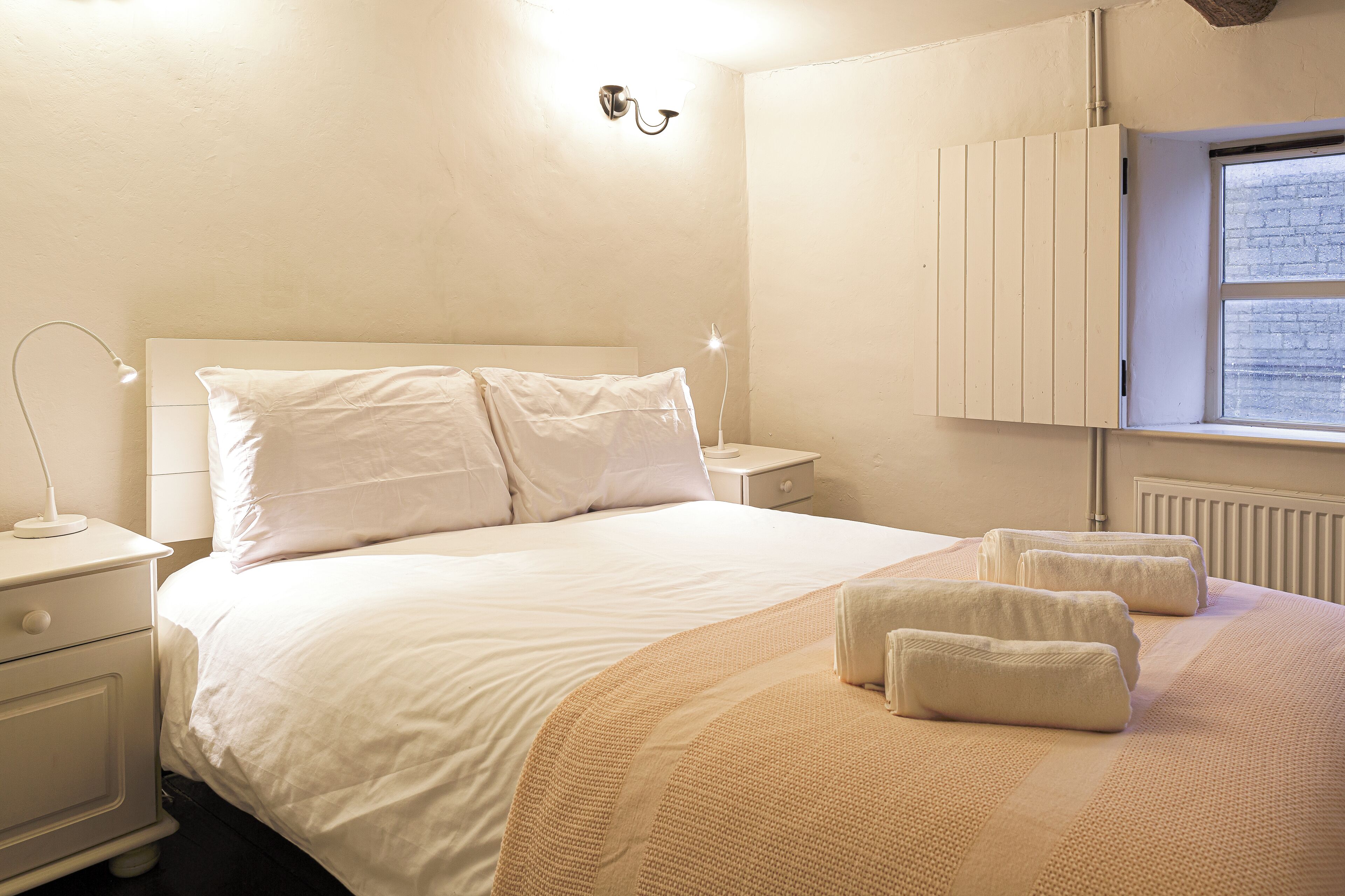Un lit bien fait, avec du linge de lit blanc et un plaid beige, une lampe de chevet et une table de chevet dotée d’un tiroir.