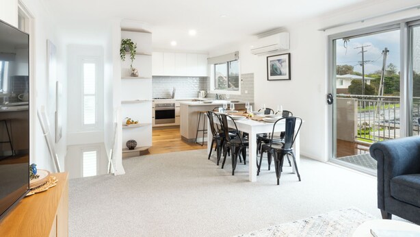 A modern kitchen with white cabinets, a dining area with a white table and black chairs, and a balcony with a view of the neighborhood. - Creek House (Ocean Grove)