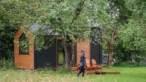 A cabin with a sloped roof, a large window, and a door. There is a bench outside and a tree in front of the cabin. - Lisi Grun Tiny House by Tiny Away (Sattledt)
