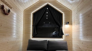A cozy cabin room with a wooden interior, a large window with curtains, a sofa, and a wall-mounted light fixture. - Lisi Grun Tiny House by Tiny Away (Sattledt)