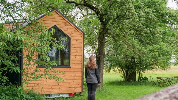 A small wooden cabin with a pitched roof and a large window, surrounded by lush greenery and trees.