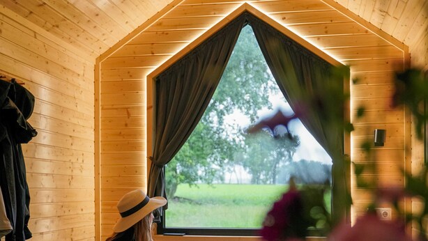 A room with wooden walls, a large window with curtains, and a view of greenery outside. - Lisi Grun Tiny House by Tiny Away (Sattledt)