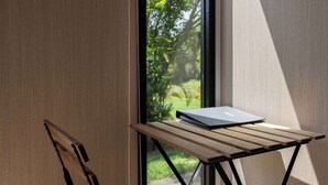 Een kamer met een houten tafel, een laptop, een raam met uitzicht op groen en een houten deur.