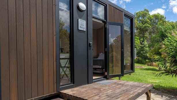 Een compact, modern vakantiehuisje met een houten gevel, een klein terras en een zichtbare slaapkamer met bed en raam.
