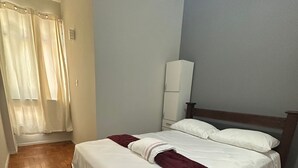 Un dormitorio con piso de madera, una cama con ropa blanca y un plaid color borgoña, un armario blanco y una ventana con cortinas de gasa.