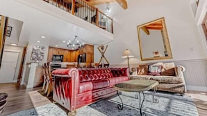 Ein geräumiges Wohnzimmer mit einem roten Samtsofa, einem runden Couchtisch und einem gemusterten Teppich. Im Hintergrund ist eine Küche erkennbar, und ein großer Spiegel ist an der Wand angebracht.