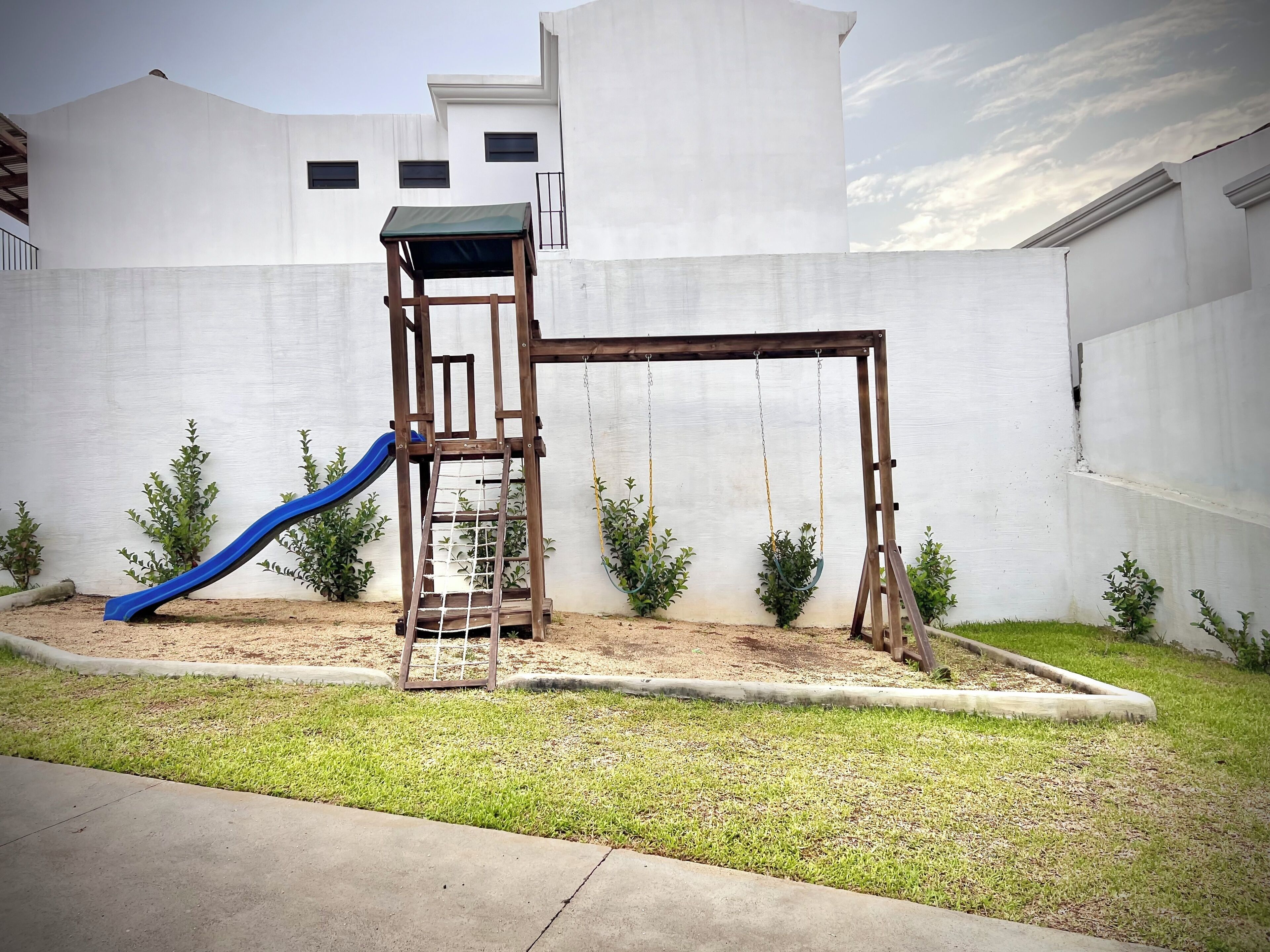 A playground with a slide and swings, surrounded by a white wall and small plants.