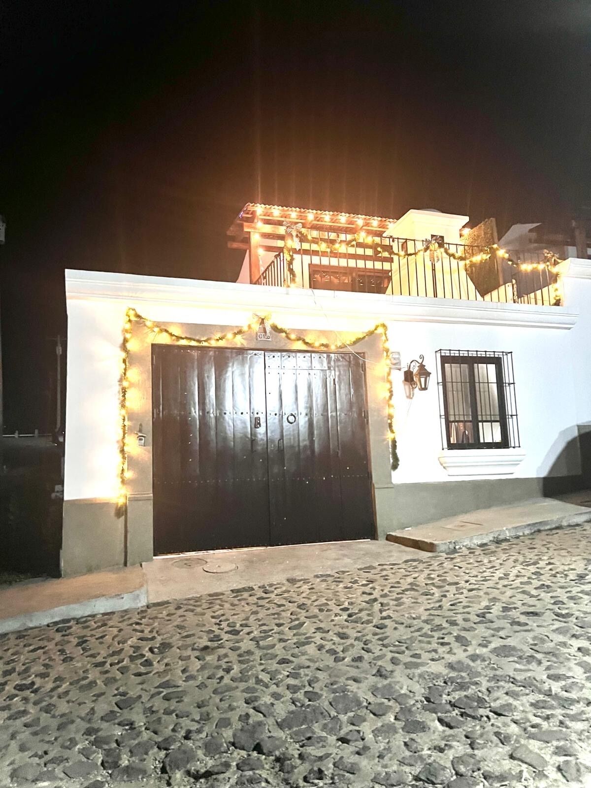 A building with a dark wooden gate, a white facade, and a balcony with string lights.