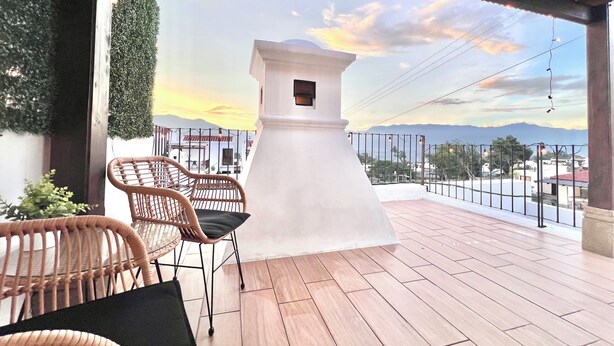 Una terraza con sillas de mimbre, un pilar blanco y vistas a edificios y montañas lejanas.