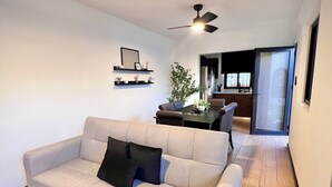 Una sala de estar con un sofá de color claro, un ventilador de techo, una zona de comedor con una mesa y sillas, y una cocina visible a través de una puerta abierta.