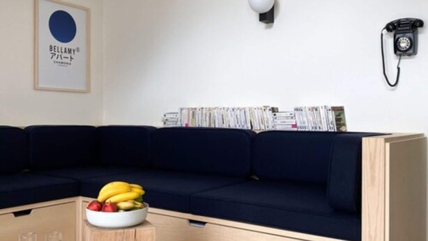 Una sala moderna con un sofá azul oscuro, una pequeña mesita de madera con un frutero, una lámpara de pared y un teléfono.