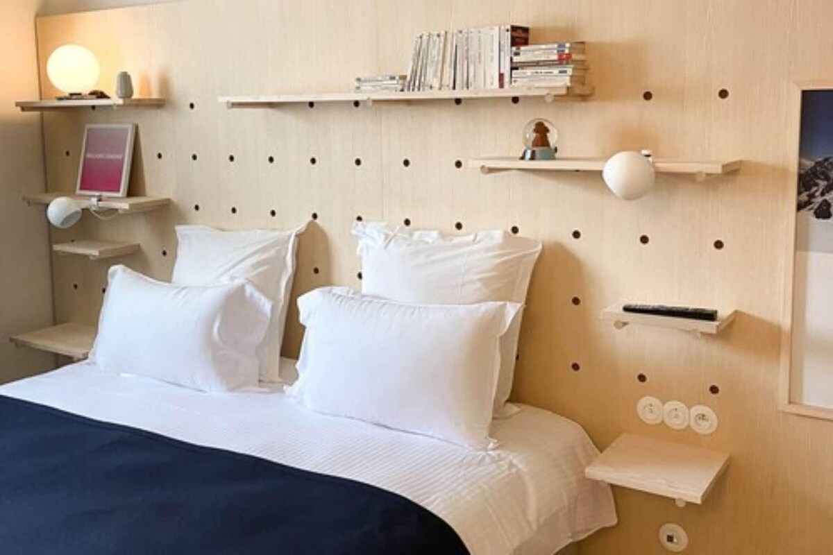 Un lit bien fait, avec du linge de lit blanc, une table de chevet avec une lampe et une étagère murale ornée de livres et d’objets décoratifs.
