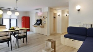 Una sala moderna con un sofá, una zona de comedor con sillas y una mesa, y una cocina con un armario rojo y una cafetera.