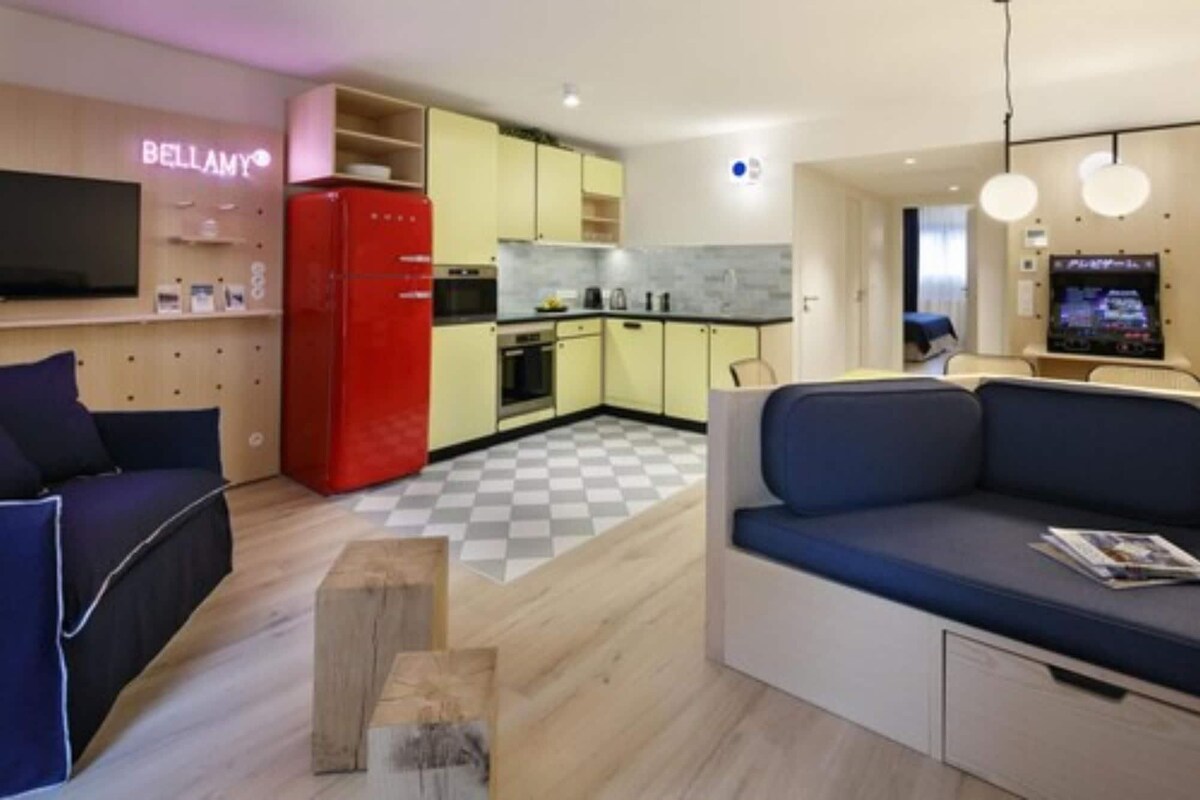 A modern living room with a sofa, a red refrigerator, and a kitchen area with yellow cabinets.