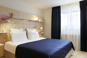 A hotel room with a bed, bedside tables, a wall-mounted shelf with books, and a window with curtains. - Appartements Bellamy (Chamonix-Mont-Blanc)