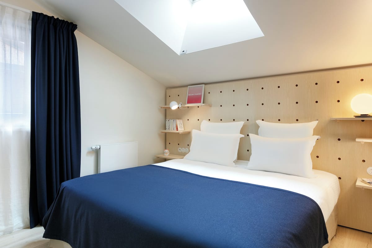 A hotel room with a bed, a bedside table with a lamp, a small shelf with books, and a window with curtains.