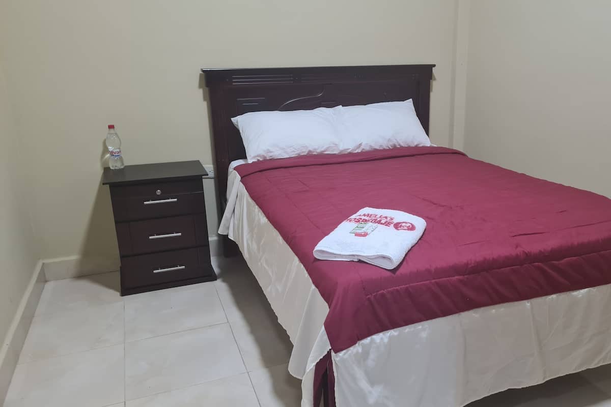 Una cama bien hecha con una almohada blanca y una manta roja, una mesita de noche de madera oscura con una botella de agua y un piso de baldosas blancas.