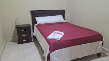 Una cama bien hecha con una almohada blanca y una manta roja, una mesita de noche de madera oscura con una botella de agua y un piso de baldosas blancas.