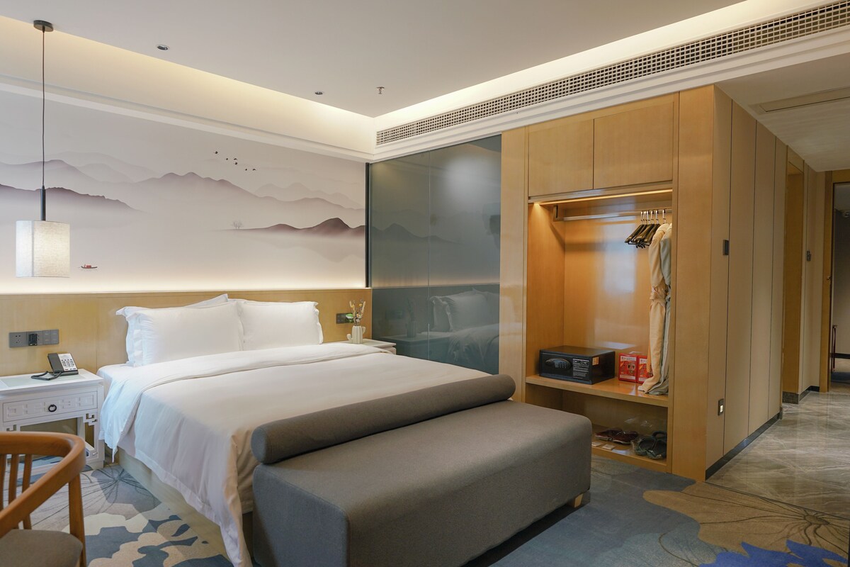Ein modernes Hotelzimmer mit einem großen Bett, einer Sitzbank, einem Kleiderschrank, einem Schreibtisch mit Telefon und einer Wand mit Bergmotiv.