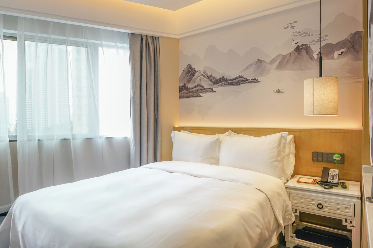 Ein Hotelzimmer mit einem großen Bett, einem Nachttisch mit einer Lampe und einer Berglandschaft als Wandgemälde.